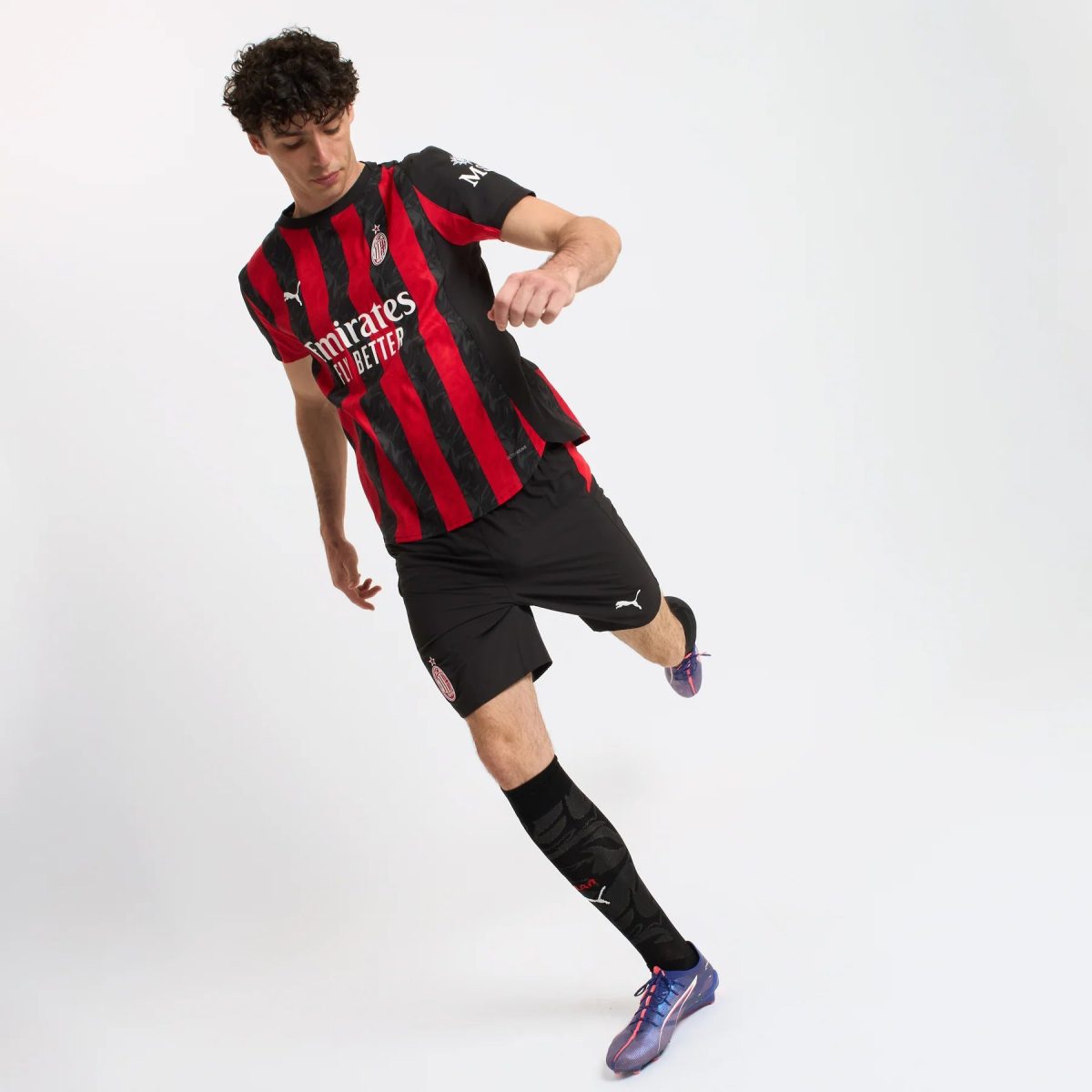 Male soccer player in a red-and-black striped kit is mid-air, kicking with his right leg against a plain white background (Emirates sponsor logo visible).