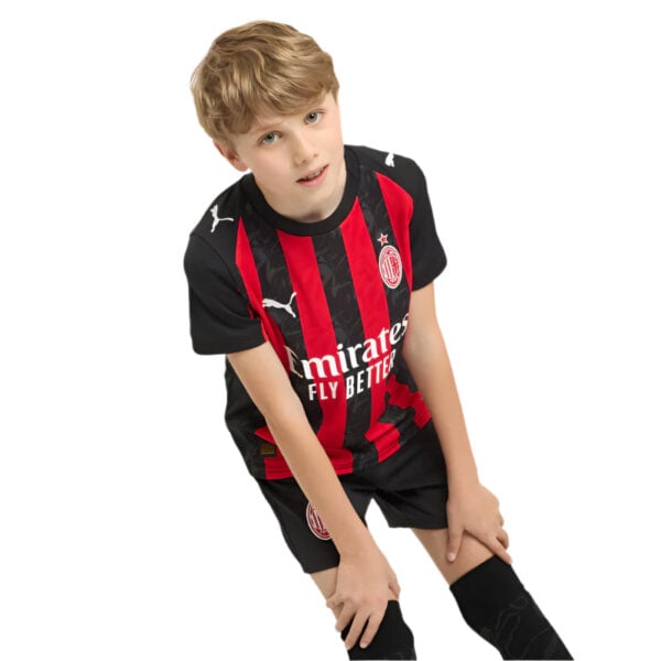 Young boy in an AC Milan soccer jersey (red and black stripes) bending forward with hands on his knees, looking at the camera.
