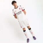 Male model wearing an Arsenal white away kit (Emirates Fly Better) with white shorts, long white socks, and blue football boots, posing with arms behind back.