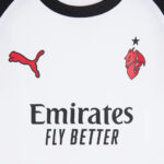 White jersey front with black collar, red Puma logo left and a red club crest with a star on the right, and 'Emirates FLY BETTER' text.