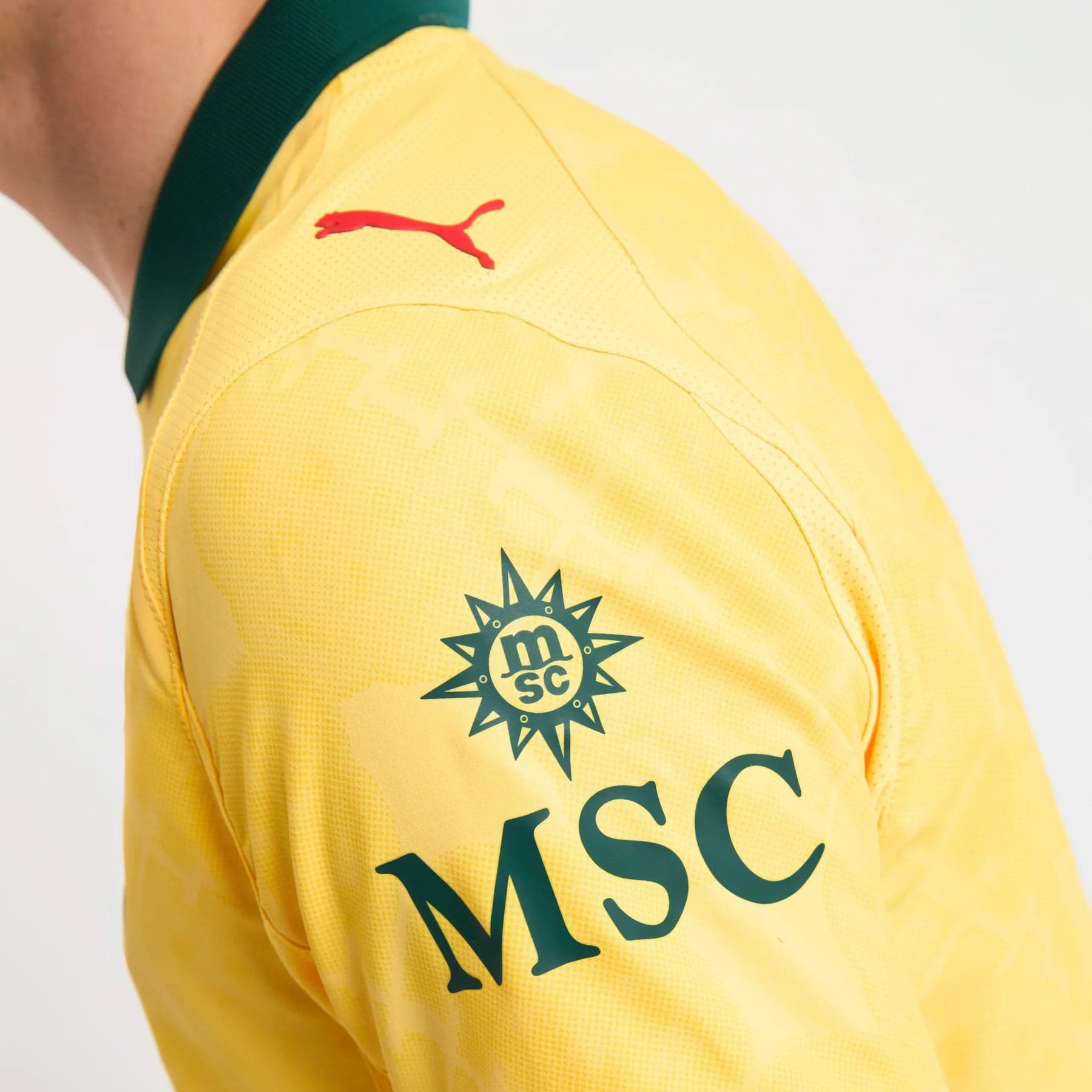 779978-C22_07_1800x1800_c6580063-0b33-459f-a69b-f51cecd3d4e8 | UAE SPORT STORE Close-up of a yellow sports jersey shoulder with a red Puma logo and a green collar trim, featuring an MSC emblem on the chest.