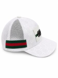 White Gucci baseball cap with a monogram pattern, mesh back, and green-red stripe strap with a metal badge on the front panel.