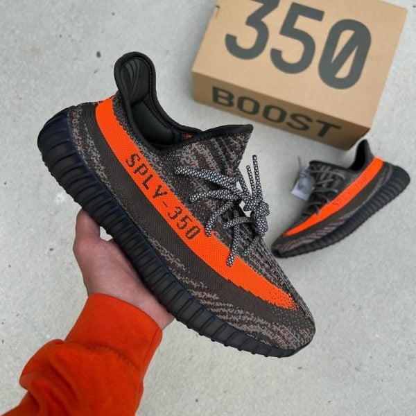 Pair of dark gray and orange Adidas Yeezy Boost 350 sneakers, one being held up with a beige 350 box in the background.
