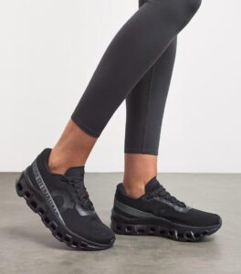 Black athletic sneakers with a chunky hollow sole, shown with black leggings on a light floor; side view highlighting the elevated sole design.