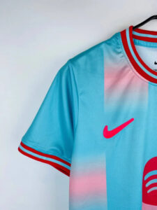 Close-up of a light blue Nike soccer jersey on a hanger, showing a pink swoosh and red-trim collar/sleeves with pink gradient panels on the torso.