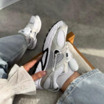 Person wearing ripped light-blue jeans sits on a concrete floor, holding a white/gray chunky sneaker with mesh panels and a bold sole.