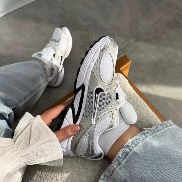 Person wearing ripped light-blue jeans sits on a concrete floor, holding a white/gray chunky sneaker with mesh panels and a bold sole.