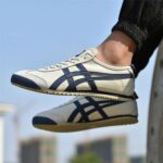 White Onitsuka Tiger sneakers with dark blue stripes, worn while jumping outdoors against a blue sky.