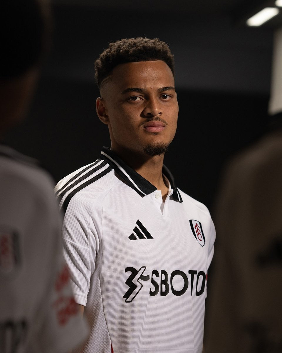 7BGkXMW4MvIpZEc | UAE SPORT STORE Portrait of a young man in a white Fulham football jersey, facing the camera in a dim backstage setting.