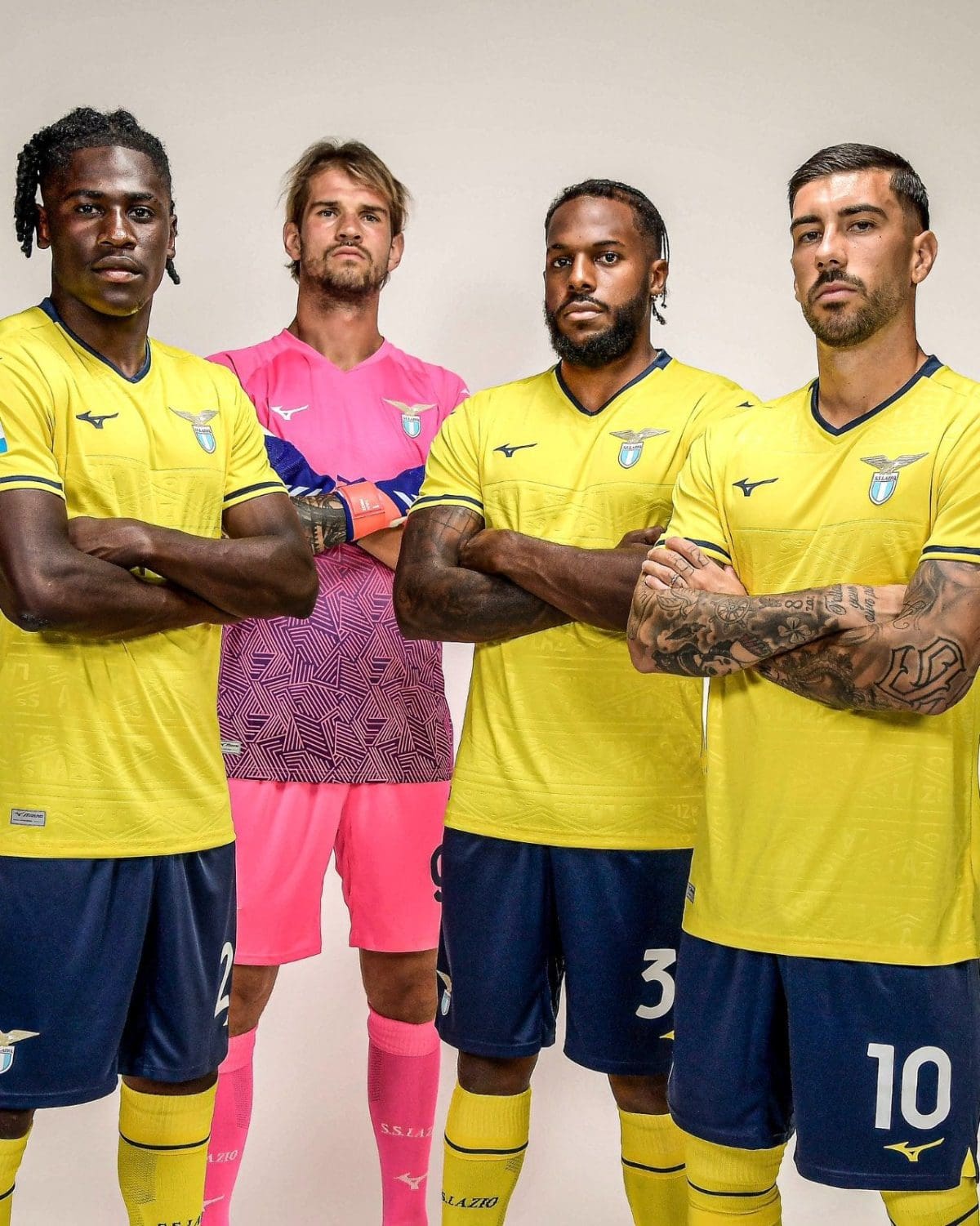 Four male soccer players stand with arms crossed, wearing yellow jerseys (one in pink goalkeeper kit) for a team photo.