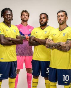 Four male soccer players stand with arms crossed, wearing yellow jerseys (one in pink goalkeeper kit) for a team photo.