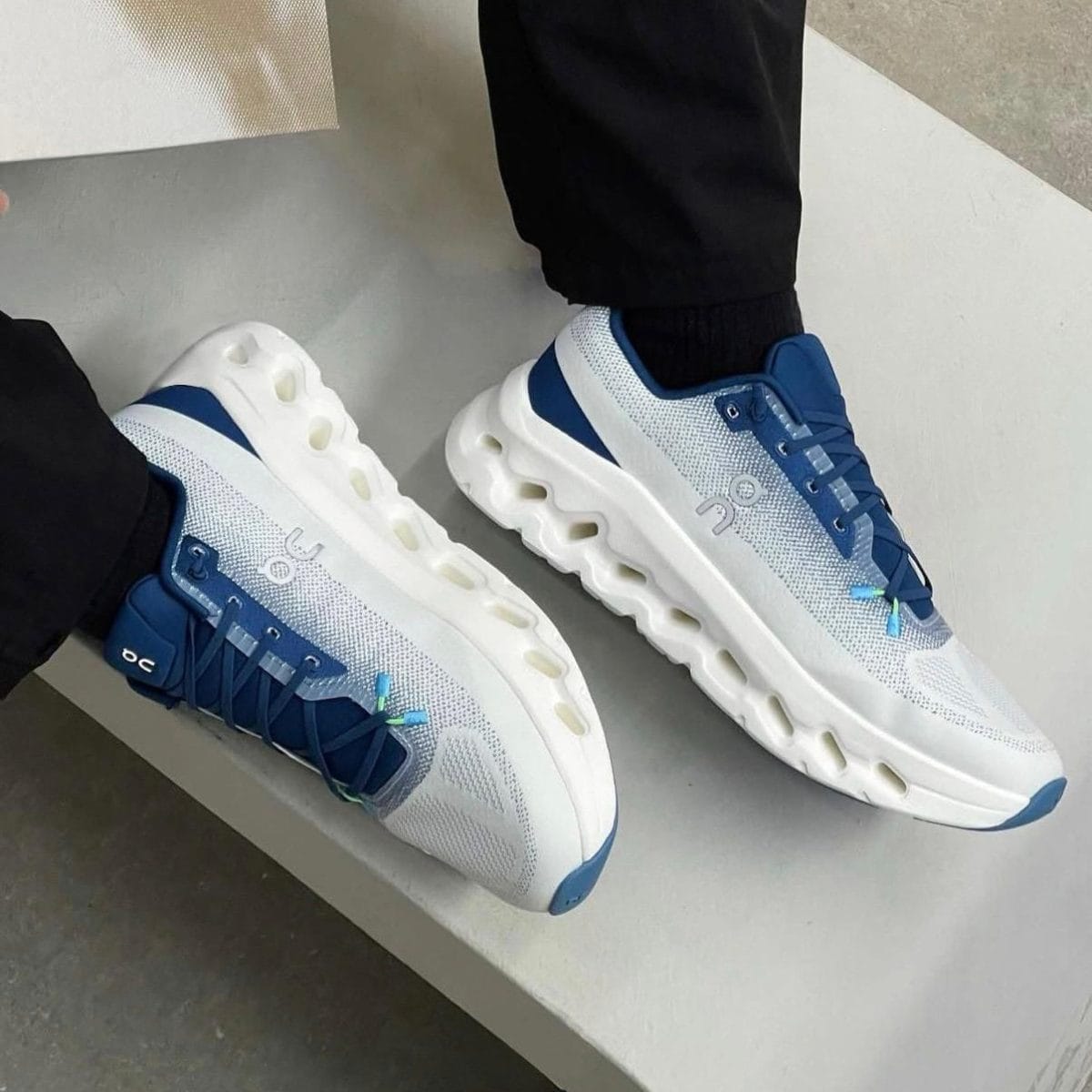 White and blue On running sneakers with a segmented hollow midsole, worn by a person in black pants.