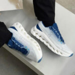 White and blue On running sneakers with a segmented hollow midsole, worn by a person in black pants.