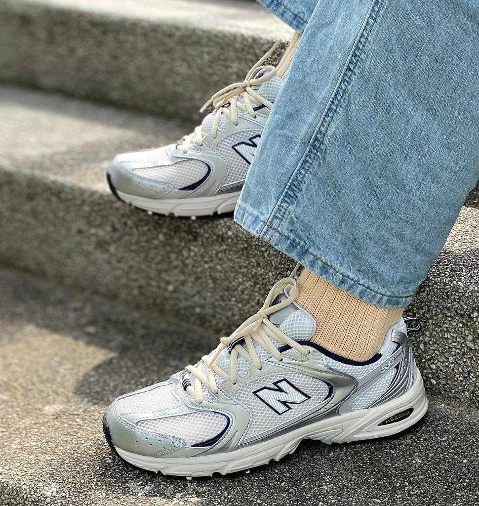 7a8f7539-3157-49ff-aec5-22a49a93bf22 | UAE SPORT STORE Close-up of light gray New Balance sneakers on concrete steps, with beige socks and denim jeans visible.