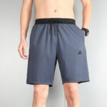 Men's gray-blue athletic shorts with black drawstring waistband, Adidas logo on the left leg, model standing with one hand in pocket.
