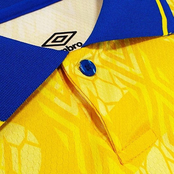 Close-up of a blue polo shirt collar over a yellow patterned shirt, with a blue button visible and the Umbro logo peeking from the neckline.