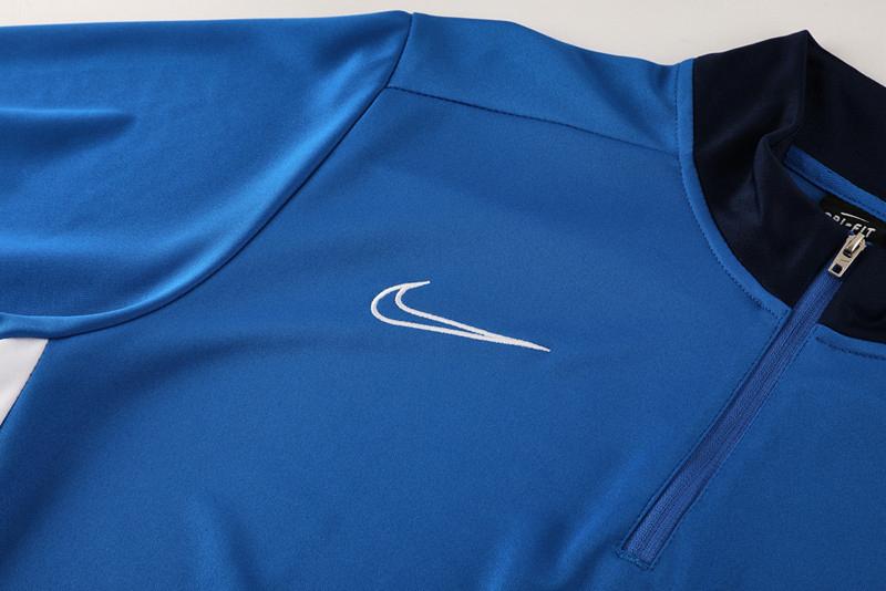 7b0e9155-edf6-4265-baeb-d6cf21dfbca2 | UAE SPORT STORE Blue quarter-zip athletic jacket with a white swoosh logo on the chest, photographed flat lay.