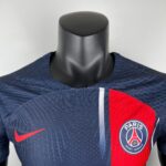 Close-up of a dark blue Paris Saint-Germain football jersey on a mannequin, showing the PSG crest and a red vertical stripe.