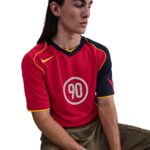 Person wearing a red sports jersey with the number 90 in a white circle, yellow trim, and navy sleeves, seated against a white background.