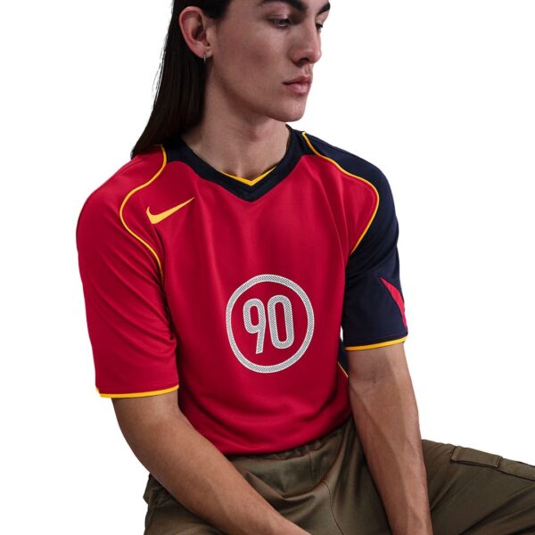 Person wearing a red sports jersey with the number 90 in a white circle, yellow trim, and navy sleeves, seated against a white background.