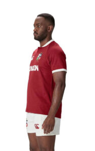 Man in profile wearing a red rugby jersey with white trim, standing in a neutral pose, crested badge on chest and white shorts with logos on the leg.