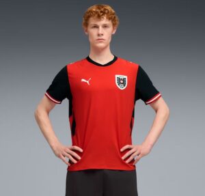 Male soccer player modeling a red and black jersey with crest, standing with hands on hips against a gray gradient background.