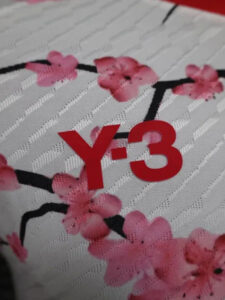 Close-up of white fabric with subtle diagonal stripes, pink flower pattern, and a bold red 'Y-3' logo.