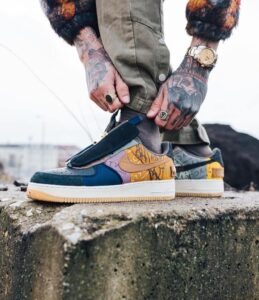 Close-up of a person tying colorful patchwork sneakers, tattoos on hands and a gold watch visible outdoors on a concrete surface.