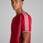 Young man wearing a red athletic jersey with white stripes on the sleeves, angled to camera, neutral background