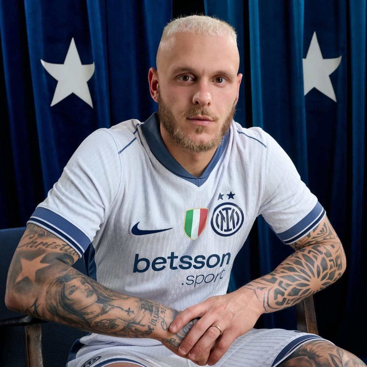 Portrait of a tattooed man with a blond buzz cut wearing a white Inter Milan jersey, seated in front of a blue curtain with white stars.