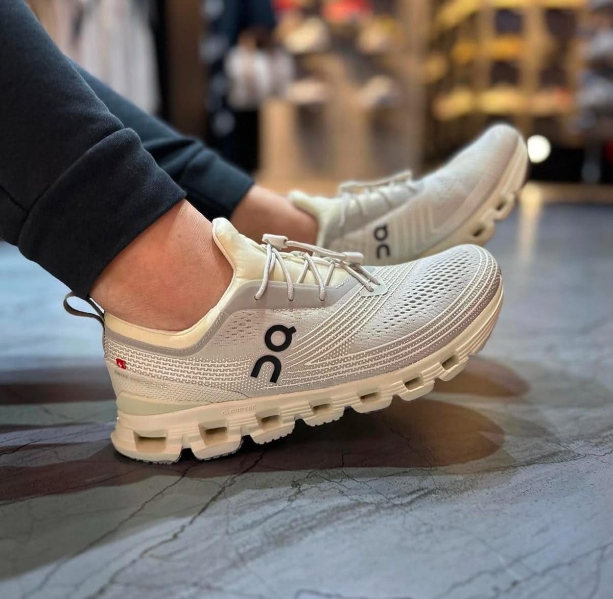 Pair of white/gray On running shoes with knit mesh, worn by a person indoors on a store floor.