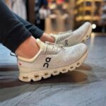Pair of white/gray On running shoes with knit mesh, worn by a person indoors on a store floor.