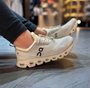 Pair of white/gray On running shoes with knit mesh, worn by a person indoors on a store floor.