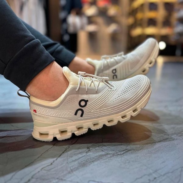 Pair of white/gray On running shoes with knit mesh, worn by a person indoors on a store floor.