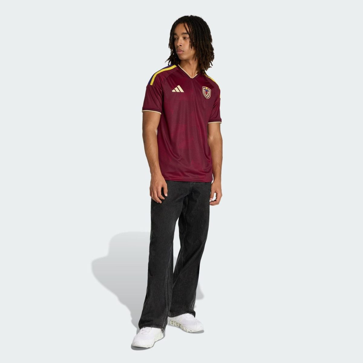 Male model wearing a maroon soccer jersey with a crest and Adidas logo, paired with dark jeans and white sneakers, standing against a light background