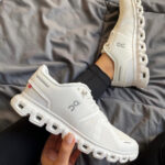 7ed704b3-0e80-4e9a-b14b-f3b1fc722cf7 | UAE SPORT STORE White On running shoes with a chunky segmented sole, one shoe resting on a bed and the other held in hand (casual indoor setting).