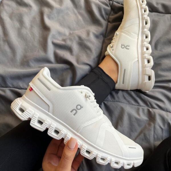 White On running shoes with a chunky segmented sole, one shoe resting on a bed and the other held in hand (casual indoor setting).