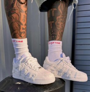 Person wearing white sneakers and white socks with a red Supreme logo, standing on a black stool, with tattooed legs visible.