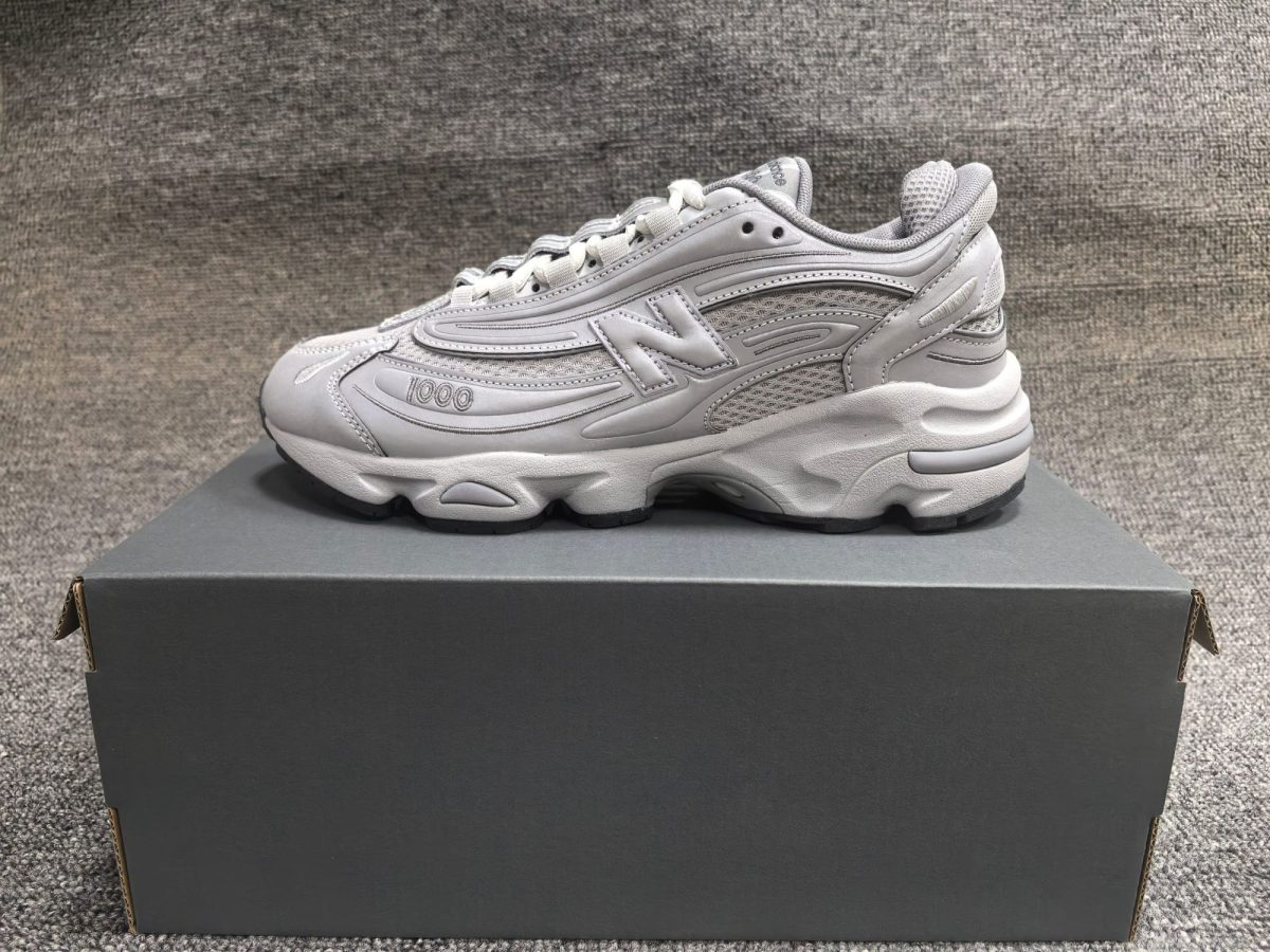 Gray athletic sneaker shown in profile resting on a dark shoe box against a textured gray carpet.