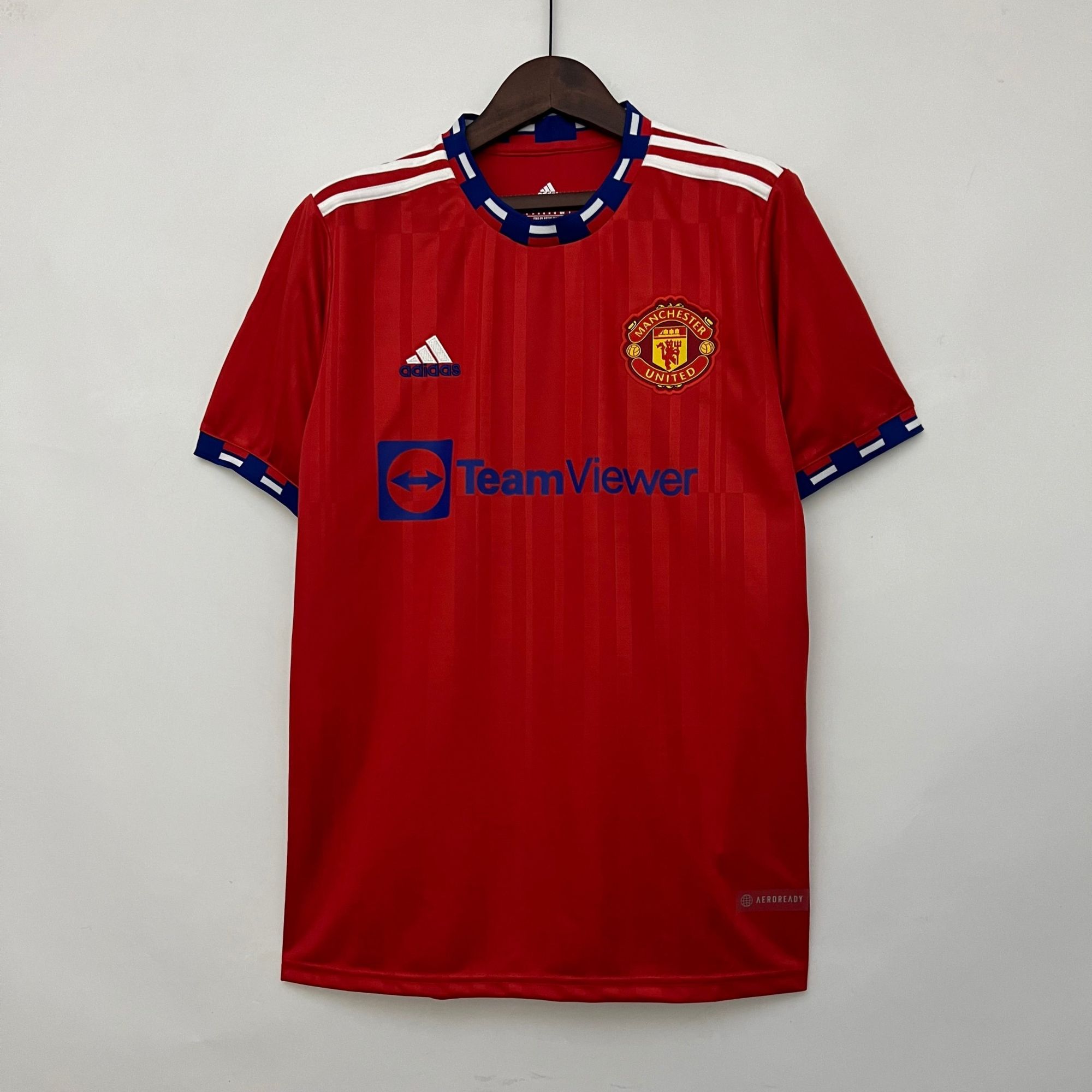 7fa5fac0_3ca472bc-c5e0-493a-ba9a-0b99a8c3ef1a | UAE SPORT STORE Red Manchester United football jersey on a wooden hanger with white stripe shoulders and blue collar, displaying TeamViewer sponsor and Adidas logo nearby.