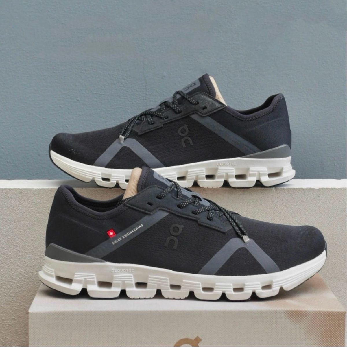 Pair of dark gray/black running shoes with thick white soles stacked on a box, showing the On logo and Swiss engineering tag.