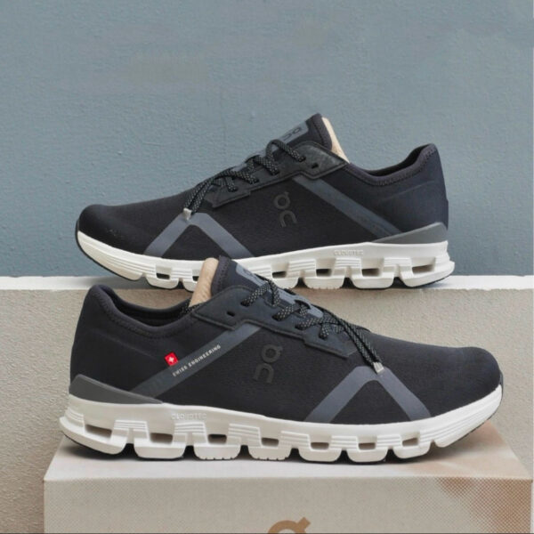 Pair of dark gray/black running shoes with thick white soles stacked on a box, showing the On logo and Swiss engineering tag.