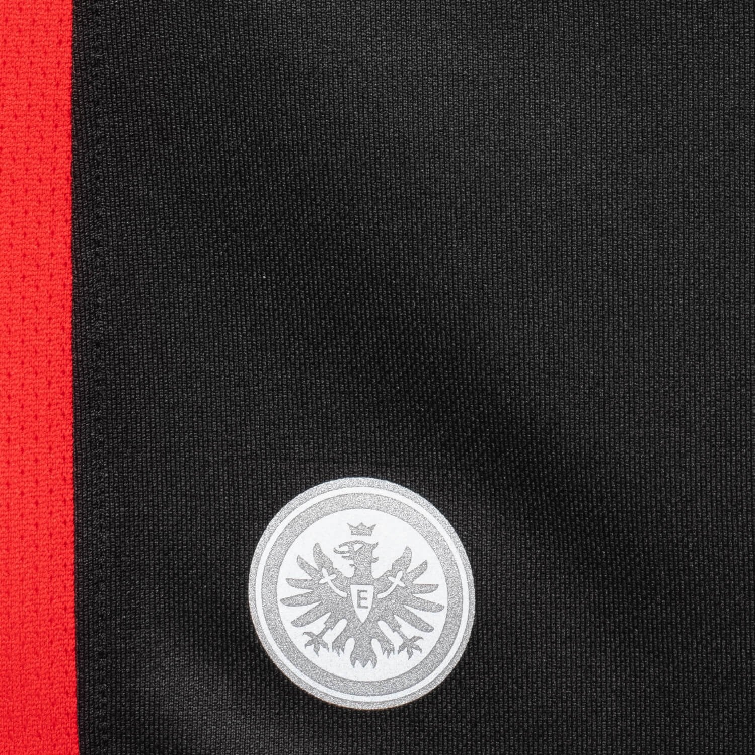 7nl6EoXakC9nJoY | UAE SPORT STORE Close-up of black fabric with a bold red vertical stripe on the left and a silver circular crest featuring an eagle in the lower center, suggesting a badge on a uniform.