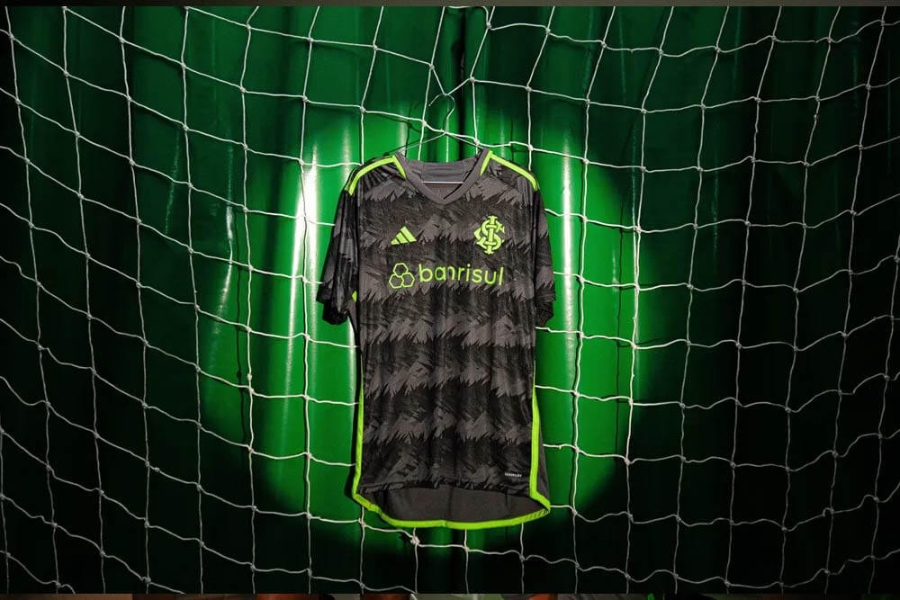 7rnUppQ9ohcpsVK_10482805-b1a7-4e26-9f29-1219684b20cc | UAE SPORT STORE Dark gray football jersey with neon green trim hanging on a goal net backdrop, sponsor banrisul and club crest visible.