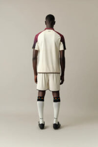 Back view of a man wearing a cream-colored soccer jersey with maroon sleeves, matching shorts, white socks, and black cleats.