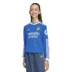 Young woman modeling a blue Real Madrid jersey with Emirates Fly Better sponsor on a white background, long sleeves with white stripes.