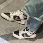 White, black, and beige Nike sneakers on blue jeans, standing on grey speckled pavement.