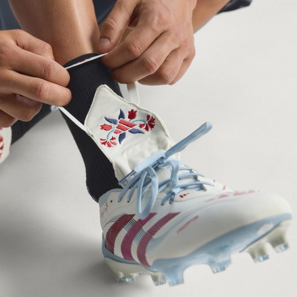 Close-up of hands tying a decorative white athletic shoe with light blue laces and embroidered red/blue motifs, worn with a black sock