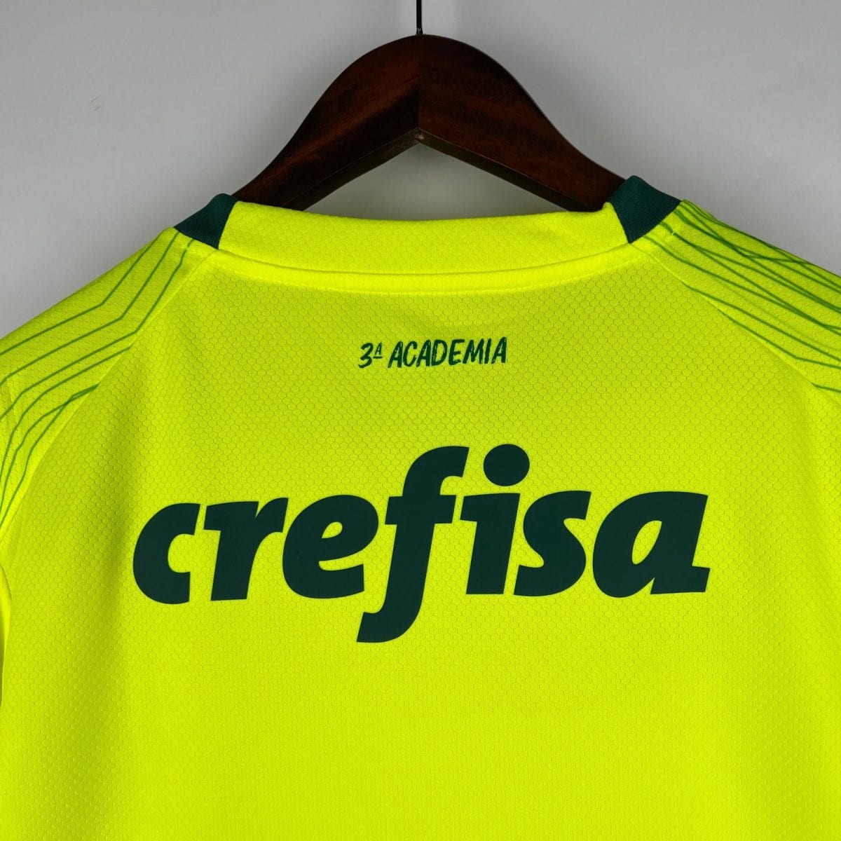 High-visibility yellow sports jersey on a wooden hanger, with dark green shoulder patches and the text 3ª ACADEMIA visible.