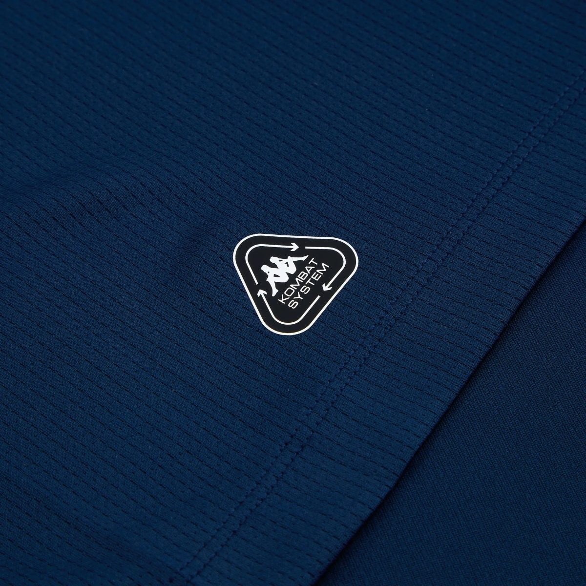 Close-up of dark blue quilted fabric with a partial black shield-shaped patch showing a white border and unreadable text at the bottom edge and stitching nearby.
